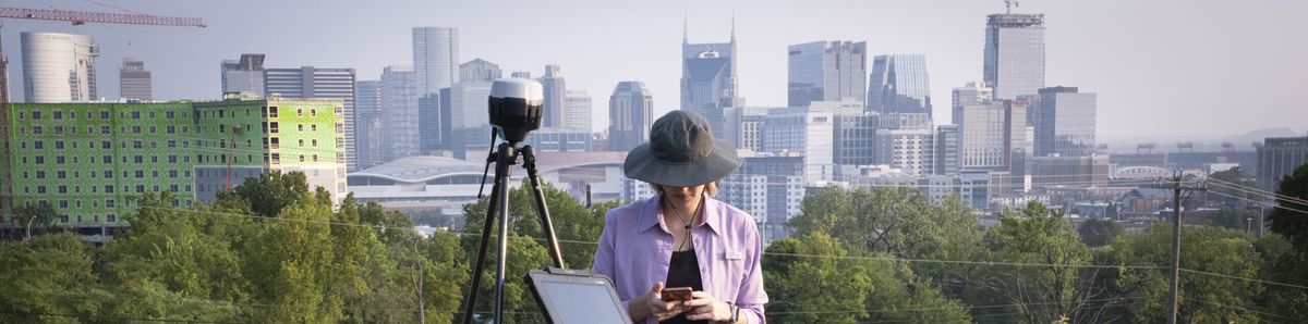 A VISR researcher records field data with a laptop and tripod-mounted survey equipment, with the Nashville skyline in the background