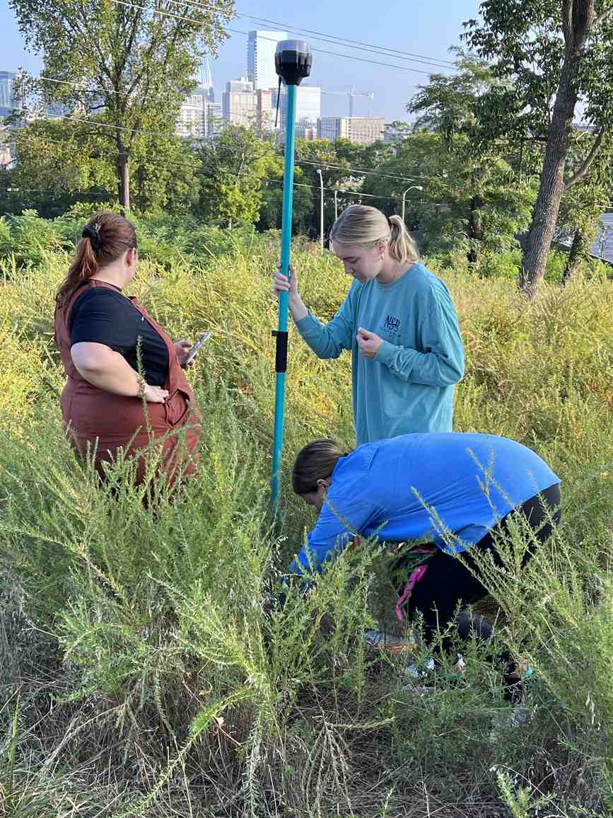 VISR researchers recording a point with an RTK GNSS rover pole in a Nashville field site, with the downtown skyline in the background