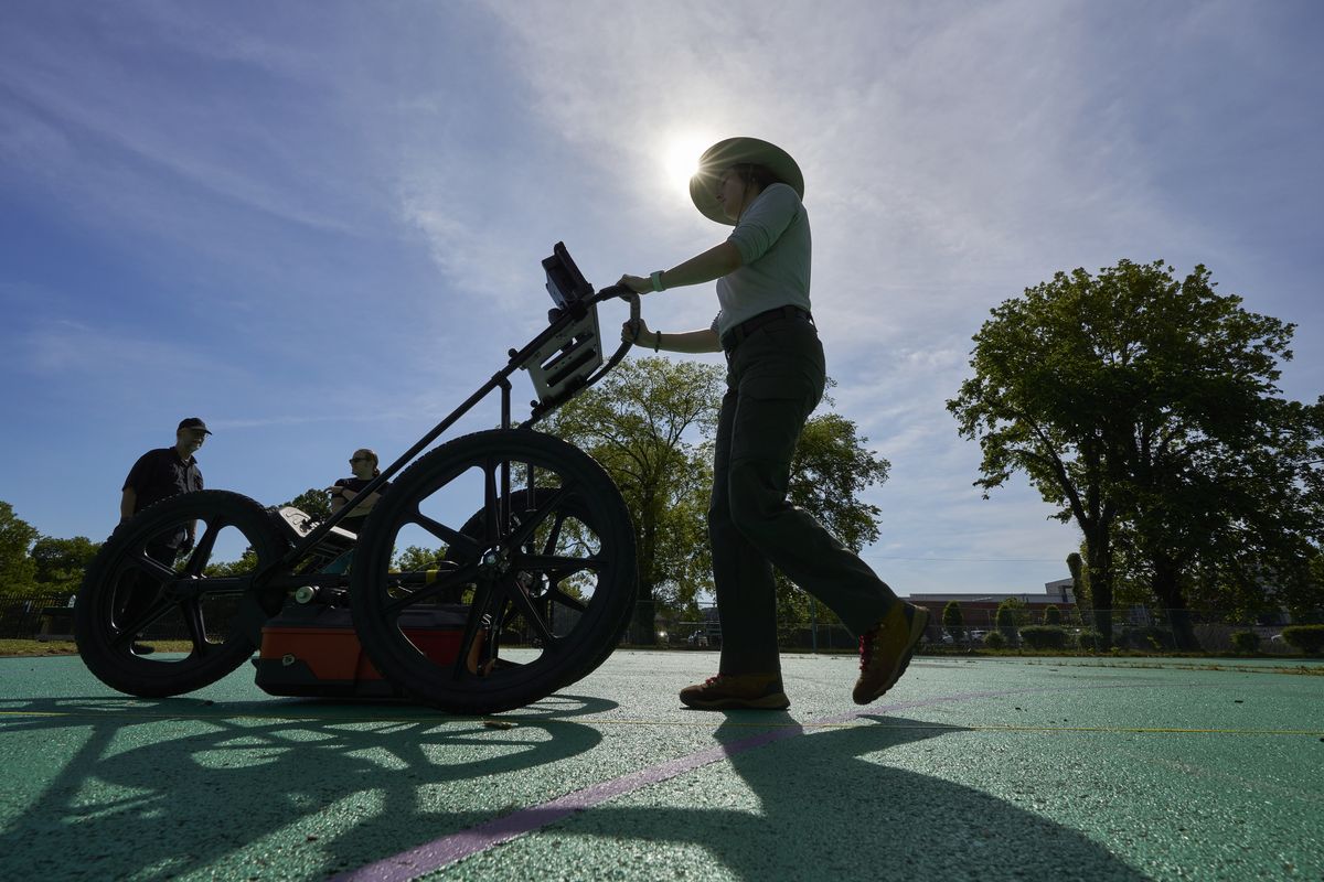 A VISR researcher pushing the GSSI UtilityScan ground penetrating radar cart across a marked survey grid, backlit by the sun, with two colleagues working behind